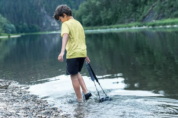 川岸にいる少年がカメラを水の向こうに隠しているいたずらな男の子の概念と防水ガジェット — ストック写真
