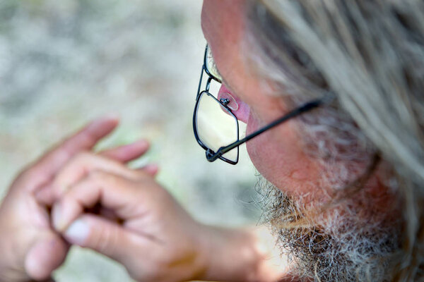 an old man with a beard and glasses looks at his hands. concept of old age and poor vision