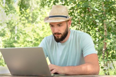 Tişörtlü sakallı bir adam dizüstü bilgisayarın başında doğa üzerine oturuyor. Uzak çalışma ve özgürlük kavramı