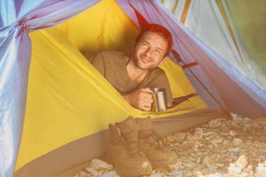 Elinde kupa olan gülümseyen bir adam çadırdan dışarı bakıyor. Yaz tatili ve turizm konsepti. ses tonlu