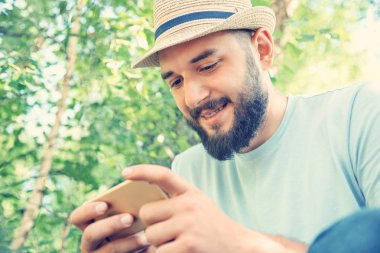 Şapkalı sakallı adam parkta oturur ve akıllı telefona bakar.