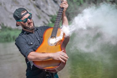 Siyah tişörtlü güneş gözlüklü bir adam doğanın arka planına karşı dumanın geldiği gitarı çalar.
