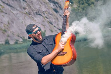 Gözlüklü sakallı bir adam ve arka planda siyah bir gömlek dumanın geldiği gitarı çalıyor.