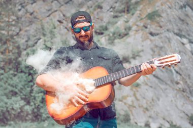 Siyah tişörtlü güneş gözlüklü bir adam doğanın arka planına karşı dumanın geldiği gitarı çalar.