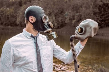 Beyaz gömlekli ve kravatlı gaz maskeli bir adam, doğanın arka planına karşı elinde bir gaz maskesi tutuyor. Enfeksiyon, kıyamet ve kirlilik kavramı