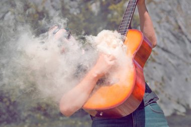 Güneş gözlüklü, kot pantolonlu bir adam doğanın arka planına karşı dumanın geldiği gitarı çalar.