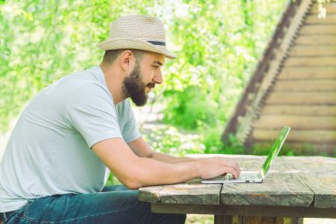 Şapkalı, sakallı bir adam doğanın arka planında bir dizüstü bilgisayarın önündeki tahta masada oturuyor. uzak çalışma ve serbest çalışma kavramı