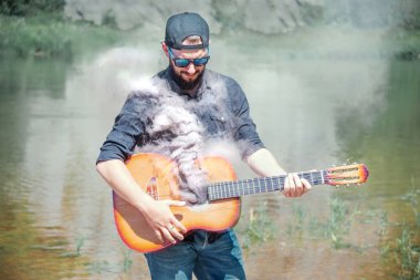 Şapkalı, güneş gözlüklü ve siyah gömlekli bir adam doğanın arka planında, elinde bir gitarla...