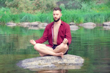 Kırmızı düğmeleri olmayan bir adam bir taşın üzerinde lotus pozisyonunda oturur ve doğada meditasyon yapar..
