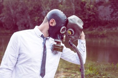 Doğanın arka planında beyaz gömlekli gaz maskeli bir adam önünde bir gaz maskesi tutuyor. Enfeksiyon, kıyamet ve kirlilik kavramları