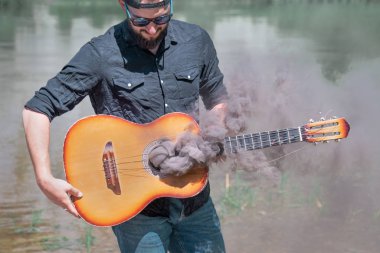 Güneş gözlüklü ve siyah gömlekli bir adam doğanın arka planında, dikkatle bir gitar tutuyor,