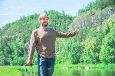 Tişört ve kot pantolon giymiş sakallı bir adam nehrin kıyısında duruyor ve oy veriyor. otostop ve seyahat konsepti