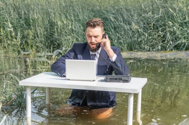 Takım elbiseli bir adam bataklıktaki beyaz bir masada pantolonsuz oturur, sabit bir telefonla konuşur ve bir dizüstü bilgisayara bakar. ofis işi kavramı