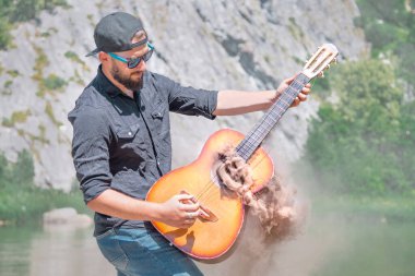Şapkalı, güneş gözlüklü ve siyah gömlekli bir adam doğanın arka planında dumanın geldiği akustik bir gitar çalar.