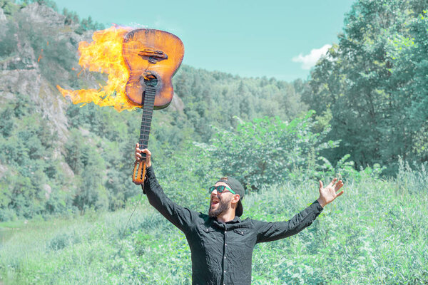 A man in a cap, sunglasses and a black shirt against the background of nature holds a burning acoustic guitar in his raised hand