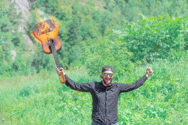 Güneşli bir yaz gününde, kepli, güneş gözlüklü ve siyah gömlekli bir adam, yükseltilmiş elinde yanan bir akustik gitar tutar.