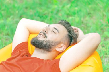 Kırmızı gömlekli sakallı bir adam sarı bir şişme yatakta yatıyor. Ellerini başının arkasına koyup yeşil çimenlerin arkasına yaslanıyor.