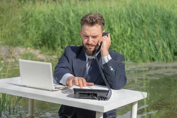 A man in a business suit sits without pants at a white table in a swamp and calls on a landline telephone. office work concept