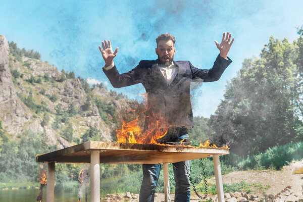 man in a business suit with hands up in a panic at a burning table against the background of nature