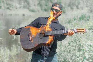 Şapkalı, güneş gözlüklü ve arka planda siyah gömlekli bir adam yanan bir akustik gitarla çalar.