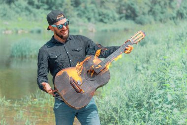 Yazın arka planında şapkalı, güneş gözlüklü ve siyah gömlekli bir adam elinde yanan bir akustik gitar tutuyor.