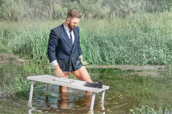 A man in a business suit without pants is standing near a white table in a swamp. office work concept