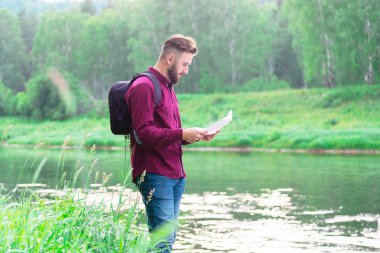 Sırt çantalı ve kot pantolonlu bir adam bölgenin haritasına bakıyor. Yazın arka planında nehir kenarındaki bir gezgin yolu belirler.