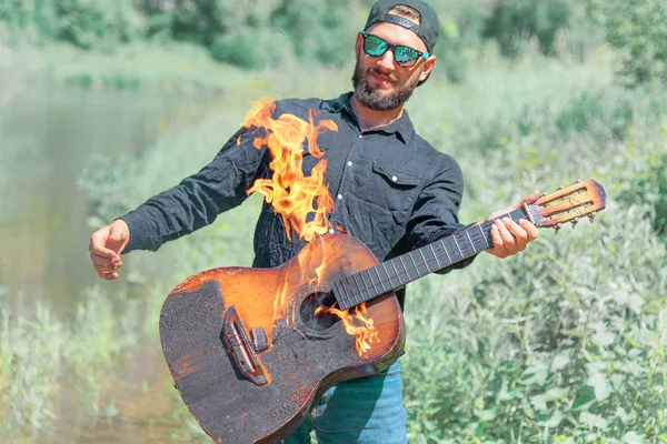 Şapkalı, güneş gözlüklü ve siyah gömlekli bir adam yanan bir gitar çalar. Yazın arka planında sakallı gitarist.