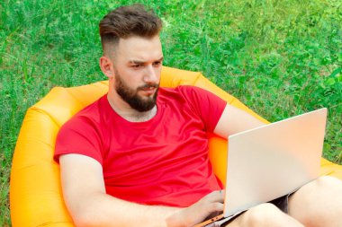 Sakallı ve kırmızı tişörtlü bir adam yaz günü şişme turuncu bir yatakta yatar ve dizüstü bilgisayarın ekranına bakar.