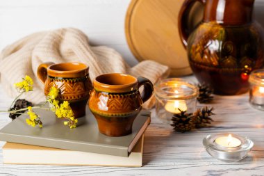 Ahşap mutfak masasında iki kaba fincanla, kitaplarla ve mumlarla sürahi kompozisyonu..