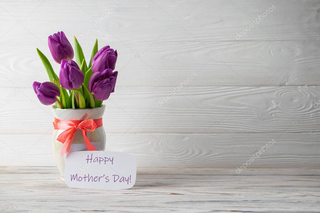 Mother's Day holiday card with a bouquet of fresh purple tulips in a vase with a pink bow on a white wooden background. Flat lay with copy space for you text