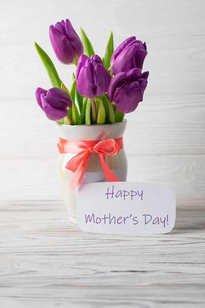 Mother's Day holiday card with a bouquet of fresh purple tulips in a vase with a pink bow on a white wooden background