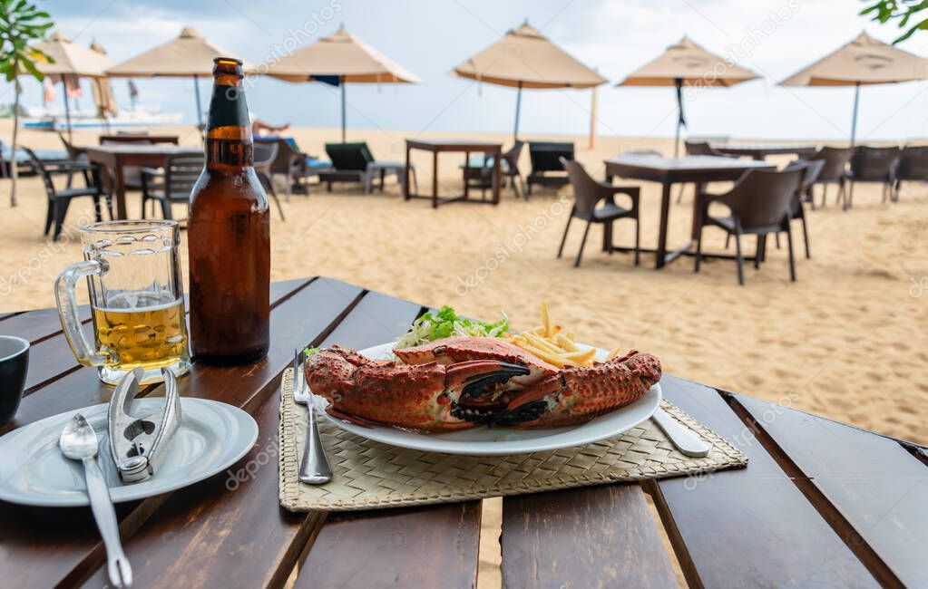 Lunch in a beach restaurant with boiled crab shell, french fries, salad on a white plate and cold light beer, sandy beach on the background.