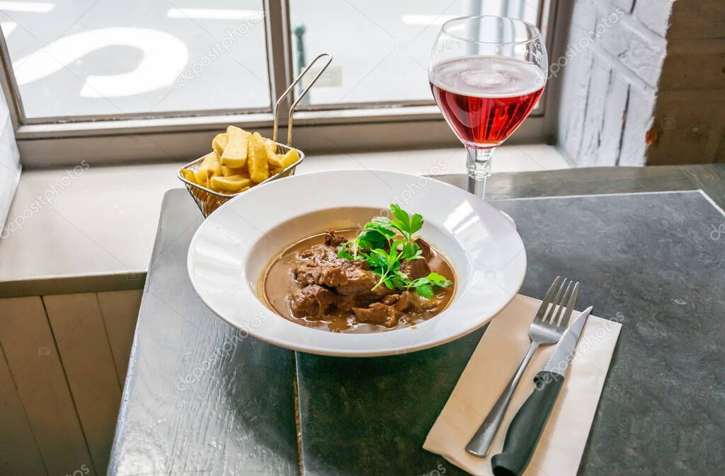 Beef stew or goulash in a delicious rich brown gravy on a white plate with french fries and glass of drink over a window background.