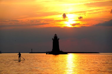 Silhouette of a light house and a man on a paddle board during sunset at Cape Henlopen State Park, Lewes, Delaware, U.S.A clipart