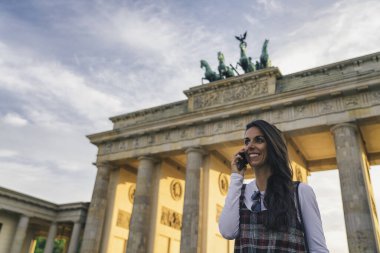Berlin, Almanya 'da Brandenburg Kapısı' nda gülümseyen havalı ve genç bir kadın cep telefonuyla konuşuyor..