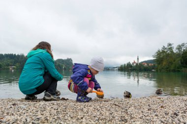 Anne ve kızı gölün kıyısında oynuyorlar. Arka planda romantik bir kilise var..