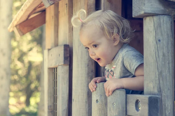 Söt blond flicka leker i trä hus tittar genom fönstret. — Stockfoto
