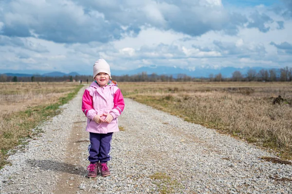 Menina bonito vestindo jaqueta rosa em pé na estrada que atravessa os campos . — Fotografia de Stock