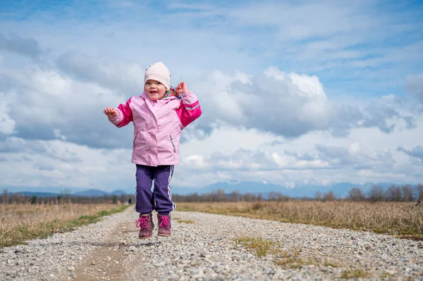 Menina bonito vestindo jaqueta rosa pulando na estrada de terra cercada de campos . — Fotografia de Stock