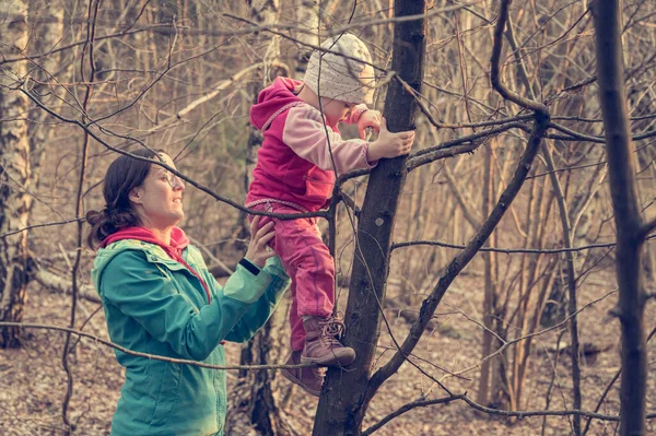 Orgulhosa mãe ativa ajudando sua filha a subir uma árvore na floresta . — Fotografia de Stock