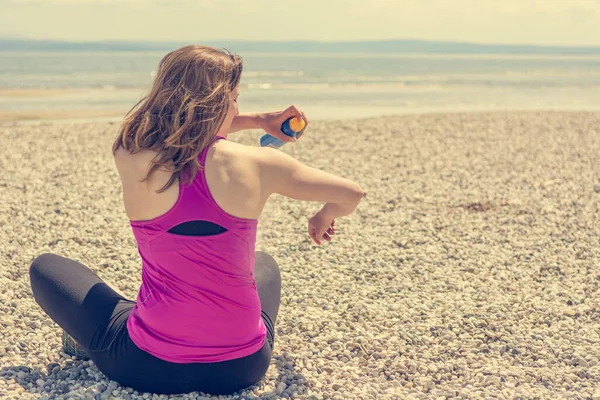 Active woman applying sunscreen spray on a pebble beach.