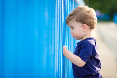 Mavi tişörtlü şirin sarışın çocuk, mavi çitten bakıyor..