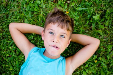Çocuk elleri başının arkasında çimenlerde yatıyor. Komik yüz ifadesi.