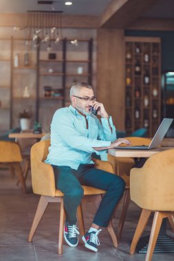 Kafede telefonla konuşurken dizüstü bilgisayarda çalışan orta yaşlı yakışıklı bir adam.