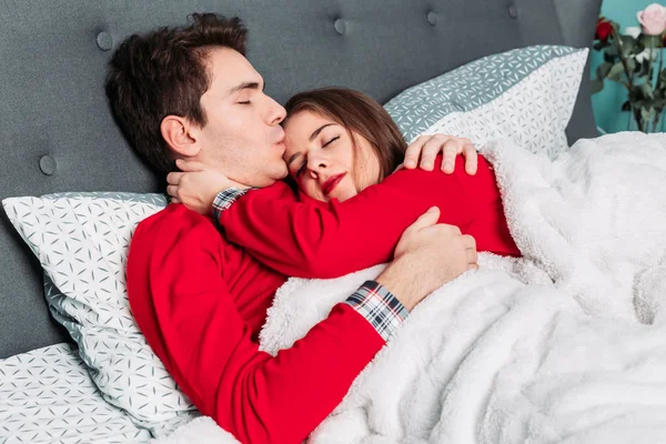 Casal deitado na cama em pijama vermelho combinando, abraçando, beijando e parecendo feliz . — Fotografia de Stock