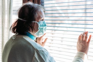Yüzünde cerrahi maske olan yaşlı bir kadının dışarıdaki pencereden endişeli bakışları. Karantina zamanı