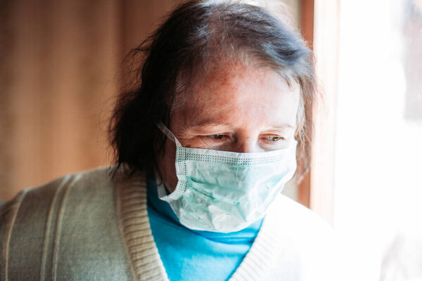 Внутреннее фото пожилой женщины с медицинской, хирургической маской на лице, выглядящей обеспокоенной. Карантинное время
