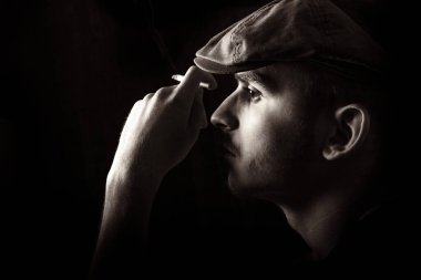 Sepia, siyah arka planda sigara içen, bıyıklı ve siyah şapkalı hoş bir gencin portresi..