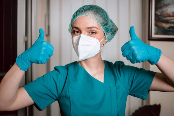 Nano maskeli, cerrahi üniformalı kadın doktor. Başparmaklarını göstererek koronavirüs salgını hakkında iyimser davranıyor. Koronavirüs salgını